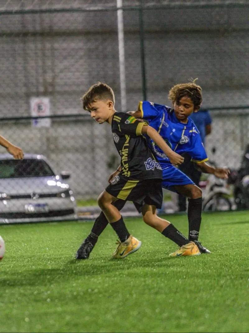 Dois atletas disputam a bola no gramado sintético, à noite